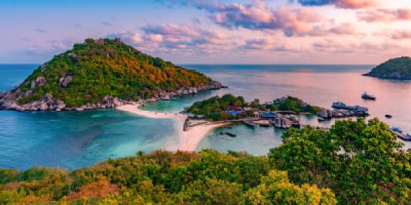 Blick auf eine grüne Insel mit Hügeln und einem kleinen Strand, umgeben von blauem Meer unter einem bunten Himmel bei Sonnenuntergang