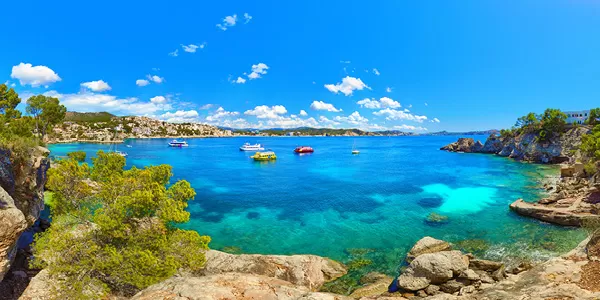 Klares blaues Wasser und Boote in einer Bucht auf Mallorca mit felsiger Küste und bewaldeten Hügeln unter blauem Himmel