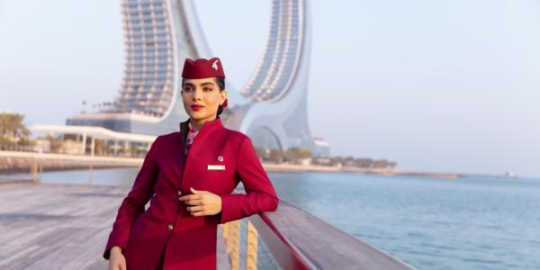 Crewmitglied bei Qatar Airways in roter Uniform steht am Geländer mit Blick auf Wasser und Stadt im Hintergrund