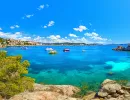 Klares blaues Wasser und Boote in einer Bucht auf Mallorca mit felsiger Küste und bewaldeten Hügeln unter blauem Himmel