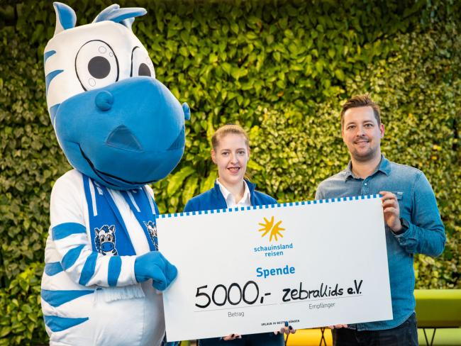 Maskottchen Hugo und Stephanie Labetzke vom Verein zebrakids nehmen von Mike Laub (schauinsland-reisen) den Spendenscheck über 5.000,- Euro entgegen.