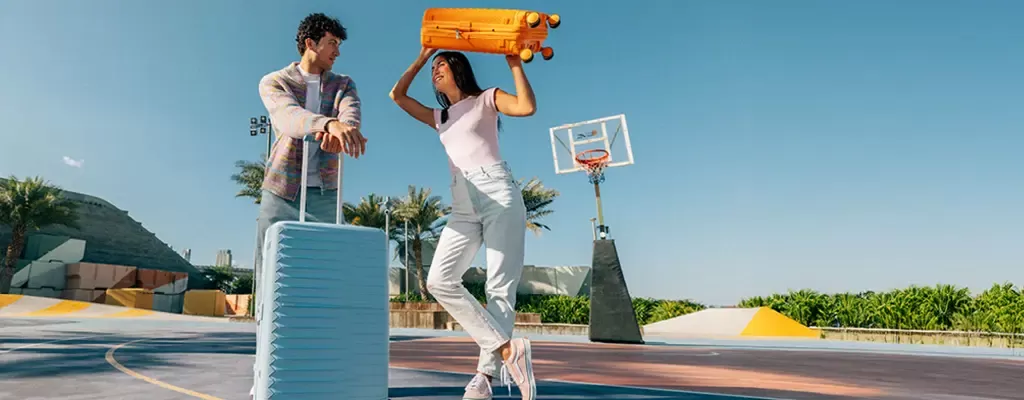 Koffer von American Tourister, Modell FastForward in Radiant Orange und Steel Blue Zwei Personen auf einem Basketballplatz, eine Frau hält einen orangefarbenen American Tourister-Koffer über dem Kopf, ein Mann lehnt an einem blauen American Tourister-Koffer